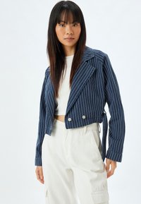 Marinrandig kort kavaj med dubbelknäppning, slagkrage, knappdetaljer och ett knytband i midjan. Vit undertröja.