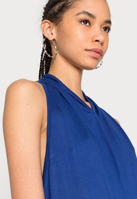 Top azul sem mangas feito de um tecido suave, com um design drapeado e decote em V. A modelo usa brincos de argola grandes.