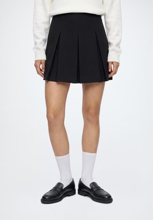 Femme portant une jupe plissée noire, un haut blanc à manches longues, des chaussettes blanches et des mocassins noirs, debout devant un fond uni.