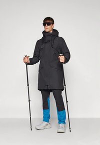 Άνδρας που φοράει μαύρο μπουφάν, μπλε και μαύρο παντελόνι, λευκά αθλητικά παπούτσια και γυαλιά ηλίου, κρατώντας δύο μπαστούνια πεζοπορίας πάνω σε απλό φόντο.