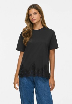Kvinde med langt hår iført en sort T-shirt med korte ærmer og blondekant samt blå jeans, der står mod en hvid baggrund.
