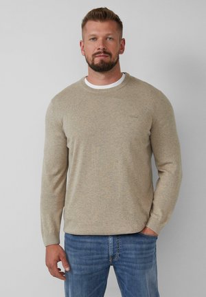 Beige gestrickter Pullover mit Rundhalsausschnitt und gerippten Bündchen, über einem weißen Hemd getragen, kombiniert mit blauen Jeans.