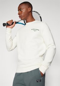 Felpa bianca con testo verde, colletto rotondo, polsini e orlo a coste. Il modello tiene una racchetta da tennis, indossando pantaloni grigi con una toppa del logo.