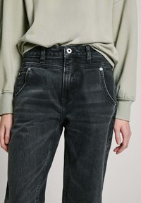 Temno sivi denim jeans s ravnim krojem, s sprednjimi žepi, gumbom za zapiranje in diskretnimi učinki cufanja. Nošeni z svetlo zelenim topom.
