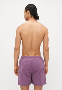 Μωβ μαγιό με ελαστική μέση και πλάγιες τσέπες, φτιαγμένο από ελαφρύ υλικό, φορεμένο από ένα άτομο από πίσω.