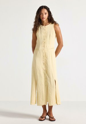 Femme aux longs cheveux bouclés portant une robe sans manches jaune clair à boutons avec des fentes à l'avant et des sandales noires, debout devant un fond uni.