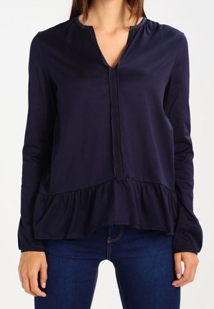 Femme portant un chemisier bleu marine foncé à manches longues avec un ourlet volanté et un col en V, associé à un jean bleu foncé.