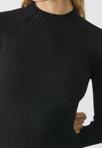 Maglietta nera a maniche lunghe con collo alto. Presenta un design testurizzato e aderente, con sottili dettagli di cucitura. Materiale morbido e elastico.