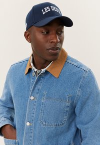 Jeansjacke mit braunem Kragen, Frontknöpfen und Brusttasche. Kombiniert mit einer marineblauen Kappe, die weißen Text "LES DEUX" trägt. Klare Textur und Farben.