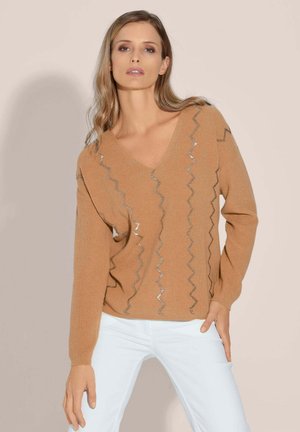 Beige V-Ausschnitt-Pullover mit langen Ärmeln und Zickzack-Silberakzenten. Strickstruktur, taillierte Passform. Kombiniert mit weißen Jeans.