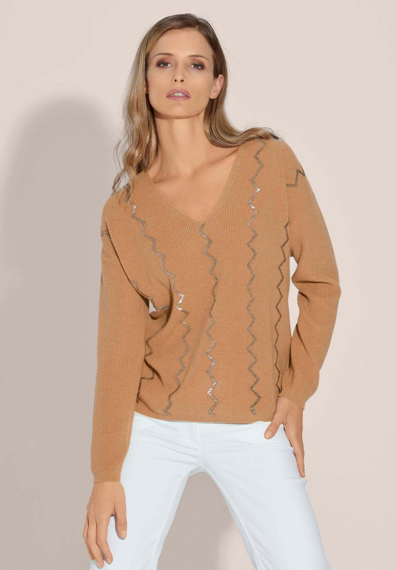 V-hals, lange mouwen trui in tan met zigzag zilveren accenten. Gebreide textuur, slim fit ontwerp. Gepaarde met witte jeans.