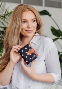 Black wallet with white polka dots and a brown leather strap. Compact design, shiny hardware, and visible logo detail on the front.
