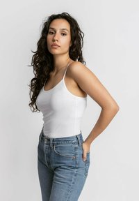 Witte katoenen tanktop met dunne bandjes, gecombineerd met hooggeknoopt blauwe spijkerbroek. Glad textuur en snug ontwerp, met minimale details.