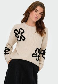 Beige knitted sweater with black floral appliqués on sleeves. Ribbed texture, round neckline, and relaxed fit. Paired with black trousers.