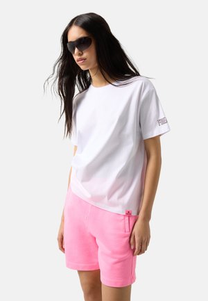 Kvinde iført sorte solbriller, hvid t-shirt med korte ærmer og tekst på ærmerne, samt lyserøde shorts med sidelommer, stående mod en hvid baggrund.