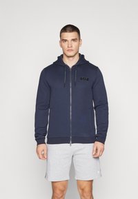 Felpa con zip blu navy in misto cotone, con toppa logo frontale, cappuccio regolabile con cordino e polsini a costine; abbinata a pantaloni shorts grigi.