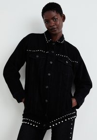 Black denim jacket with silver studs along the collar and hem. Features button closure, two front pockets, and a relaxed fit.