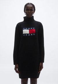 Musta neulemekko, jossa korkea poolokaulus ja resoritut hihat. Edessä logo, jossa suorakaiteen muotoinen sinivalkoinen ja punainen merkki, jossa lukee "TOMMY JEANS".