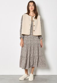Kremowa kamizelka z polaru na sukience w kwiaty z długim rękawem i plisowaną spódnicą. Całość dopełniają białe trampki.