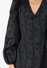 Robe noire en tissu texturé avec un motif à rayures ondulées. Manches bouffantes avec des accents transparents et un décolleté en V.