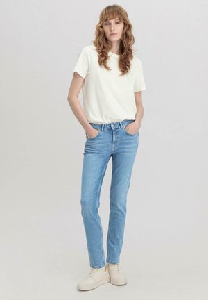 Jonge vrouw met golvend rood haar, gekleed in een wit T-shirt, lichtblauwe spijkerbroek en witte sneakers, staand met handen in de zakken tegen een effen achtergrond.