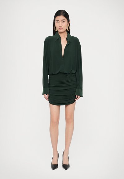 Tmavozelené šaty s hlbokým výstrihom, dlhými rukávmi a zhrnutou textúrou. Doplnene čiernymi špicatými lodičkami a zlatými kruhovými náušnicami.