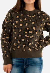 Donna che indossa un maglione leopardato marrone e beige con jeans blu, mano sui fianchi, su uno sfondo bianco.