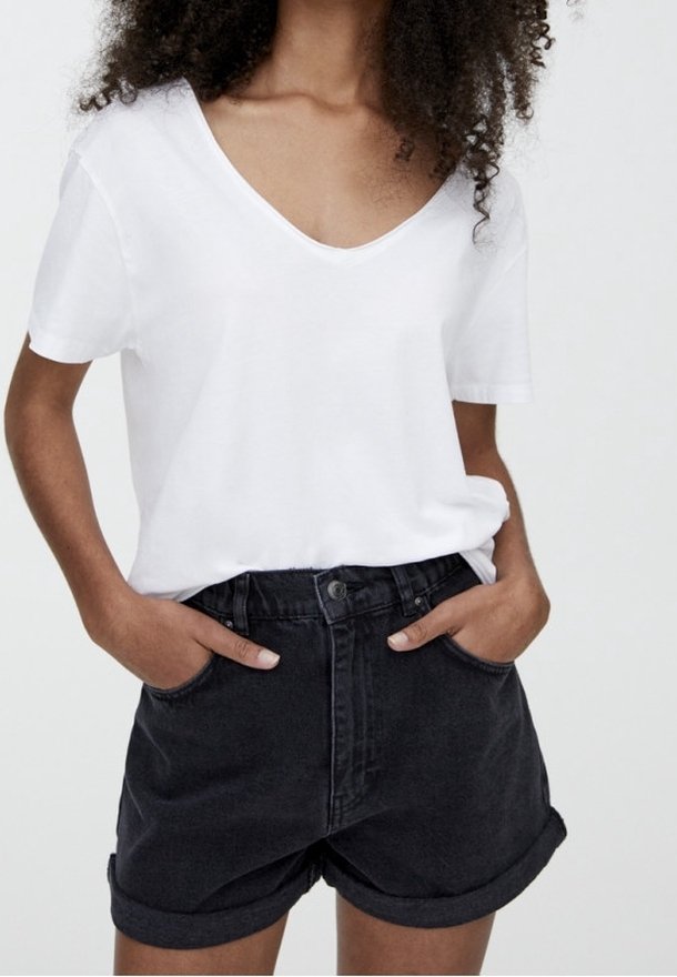 Femme portant un t-shirt blanc à col en V rentré dans un short en denim noir taille haute avec revers roulés, mains dans les poches.