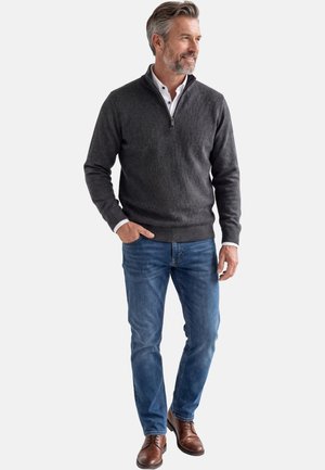 Mittelalter Mann mit grauem Haar und Bart steht lächelnd da, trägt einen dunkelgrauen Pullover mit Reißverschluss, blaue Jeans, weißes Hemd und braune Lederschuhe.