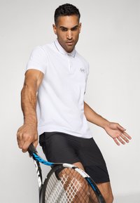 Bijela polo majica s crnim obrubom i logom na prsima, uparen s crnim sportskim šortsevima; osoba drži tenisku reketa.