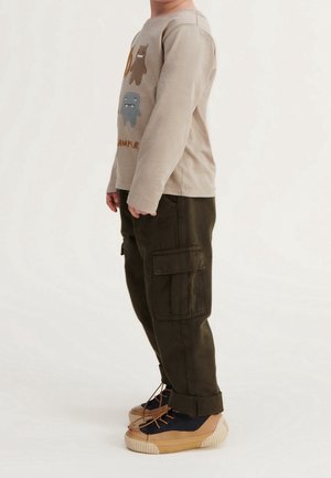 Barn iført beige langærmet skjorte med monsterprints, mørkegrønne cargobukser og tan-blå sneakers, stående i profil.