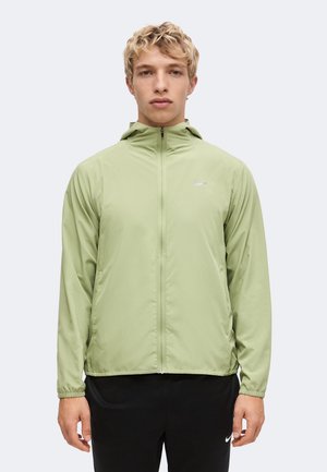 Jeune homme aux cheveux blonds et bouclés portant une veste à capuche zippée vert clair et un pantalon Nike noir sur un fond uni.