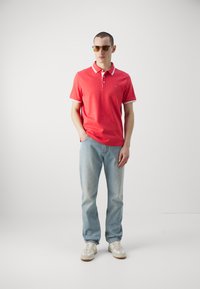 Červené polo tričko s bielymi prúžkami na golfe, skombinované so svetlomodrými džínsami a bielymi teniskami. Model nosí slnečné okuliare a stojí na jednofarebnom pozadí.