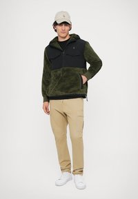 Pullover de fleece verde-oliva com parte superior em nylon preto, dois bolsos no peito e fecho zip na frente. Combinado com calças cargo bege e ténis brancos.