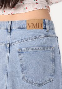 Lyseblå denimjeans med et rektangulært lærflekk i lyst brunt merket "VMD" ved midjen. Har to baklommer og synlige sømmer.