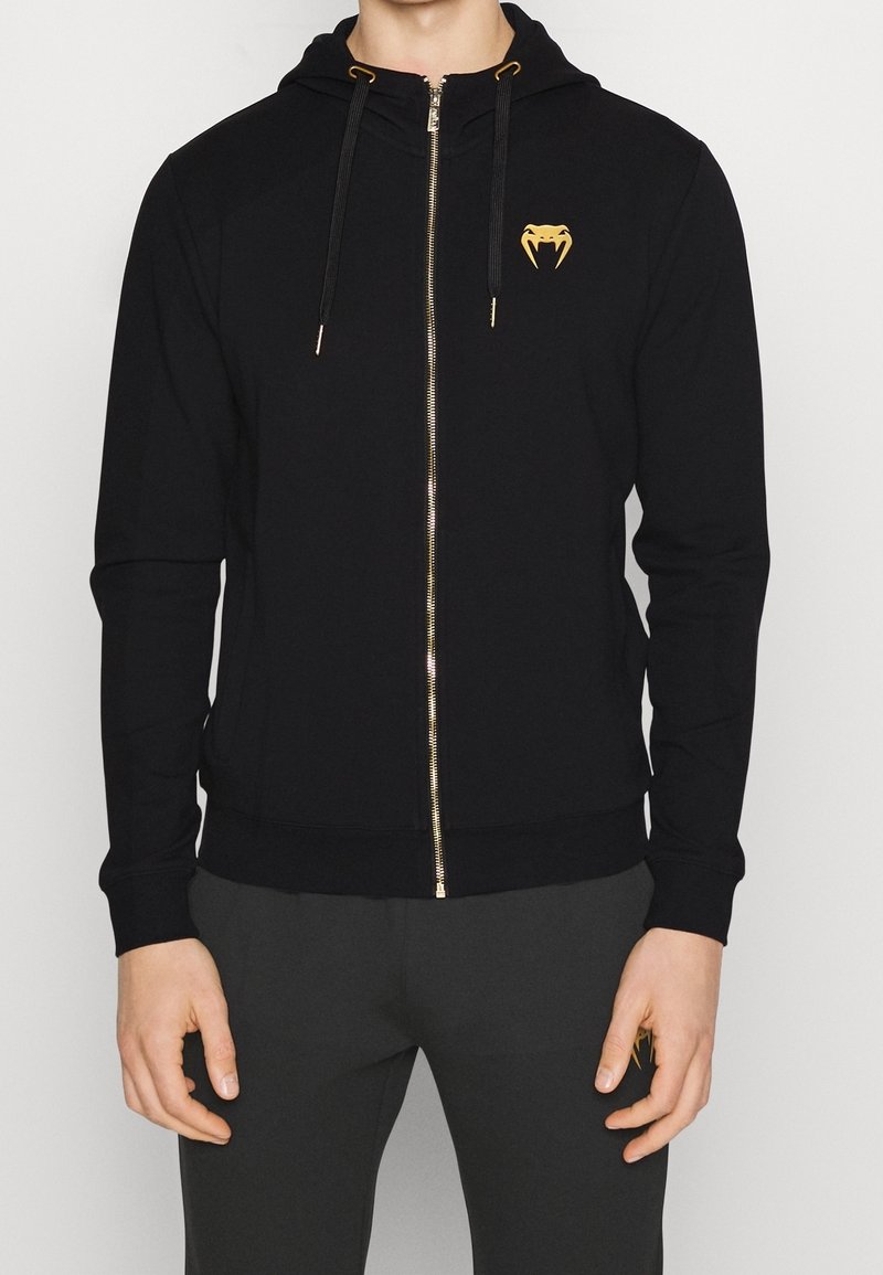 Černá mikina na zip vyrobená z měkké látky, s zlatými akcenty, kapucí a malým logem na hrudi. Model má na sobě tmavé kalhoty.