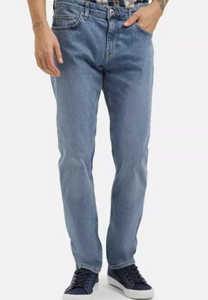 Mężczyzna ubrany w jasnoniebieskie jeansy z pięcioma kieszeniami i granatowe trampki, stoi z jedną ręką w kieszeni na białym tle.