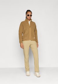 Giacca in pile marrone con chiusura a zip e tasche laterali, abbinata a pantaloni beige e scarpe chiare, posizionata su uno sfondo semplice.