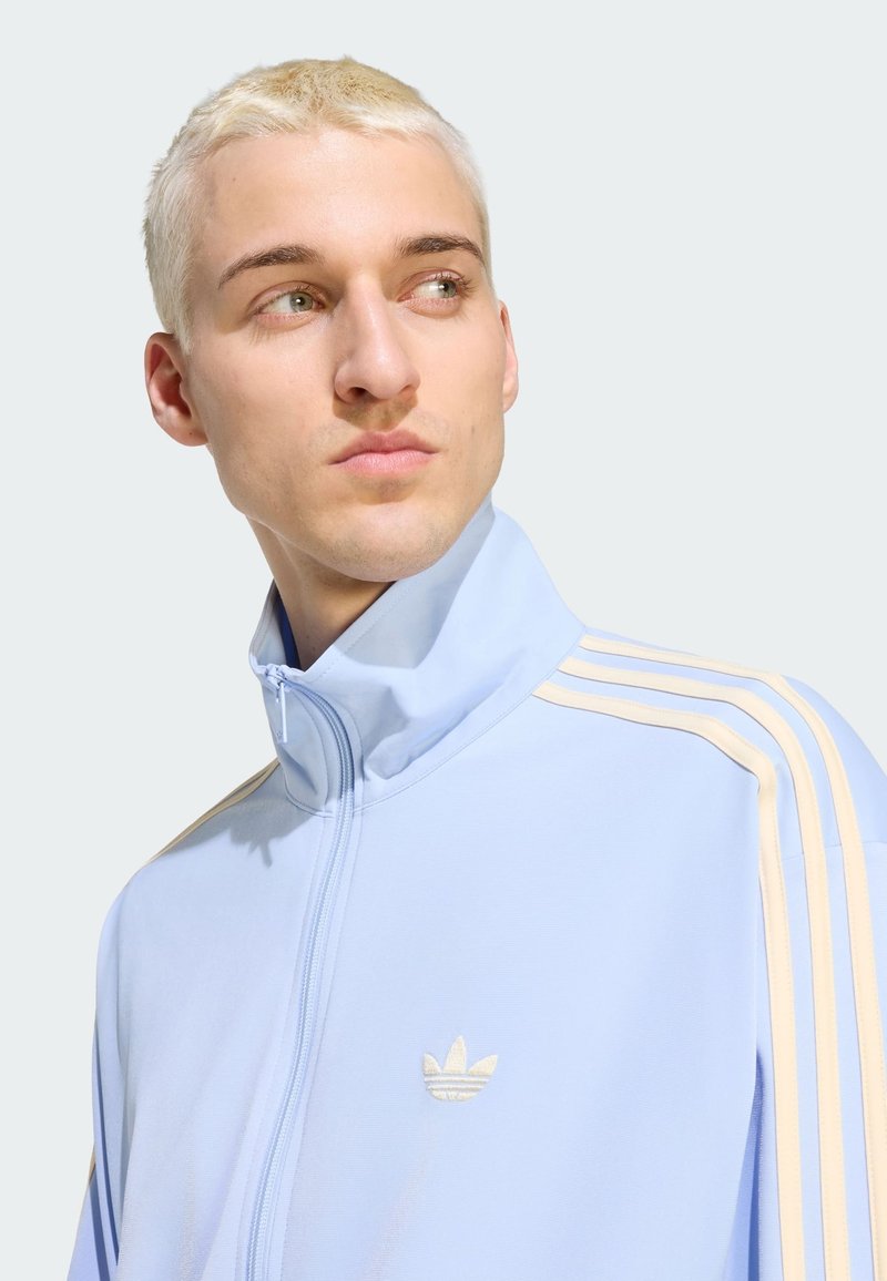 Young man with short blonde hair wearing a light blue Adidas track jacket with beige stripes, looking to the side against a neutral background.
