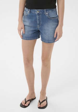 Osoba oblečená v modrých rifľových šortkách, čiernom tielku bez rukávov a čiernych žabkách stojaca pred bielym pozadím.