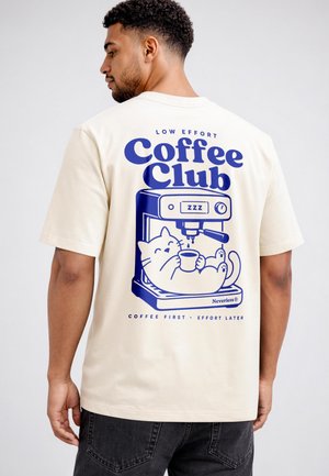 Man wearing a cream t-shirt with a blue graphic of a cat holding a coffee cup inside a coffee machine and the text "Coffee Club."