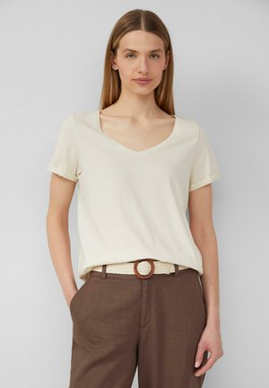 Helles Cremefarbenes V-Ausschnitt-T-Shirt mit kurzen, umgeklappten Ärmeln, kombiniert mit braunen Leinenhosen und einem geflochtenen Gürtel mit einer runden Schnalle.