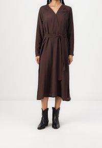 Brun omslagsklänning med långa ärmar, v-ringning och ett bälte i midjan. Tyget verkar lätt och mjukt, kombinerat med svarta anklarstövlar.