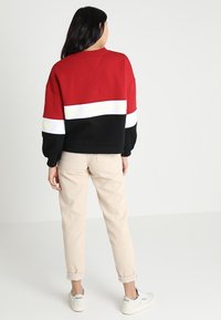 Felpa color block rossa, nera e bianca con il marchio "TOMMY". Fit rilassato, scollatura a girocollo e polsini a coste. Indossati pantaloni beige e scarpe bianche.