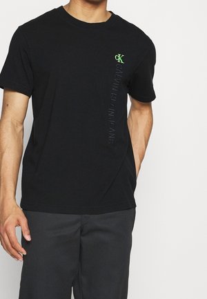 Mężczyzna w czarnej koszulce Calvin Klein Jeans z pionowym logo i neonowo zielonym emblematem "cK" na klatce piersiowej, w ciemnych spodniach.