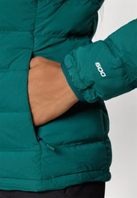Teal isolert jakke har en glidelåslomme og elastiske cuff. Merkverdig detalj: "600" trykt på cuffen. Glatt tekstur, lett design.