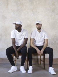 To menn sitter på trestoler mot en teksturert, lysfarget vegg. Begge har på seg hvite poloer, svarte bukser og hvite sneakers, samt matchende capser.