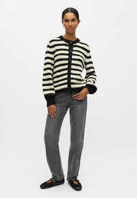 Sort og hvid stribet cardigan med knapper, tekstureret strik, og sort ribstrik ved ærmerne, parret med grå klokke jeans og sorte sko.