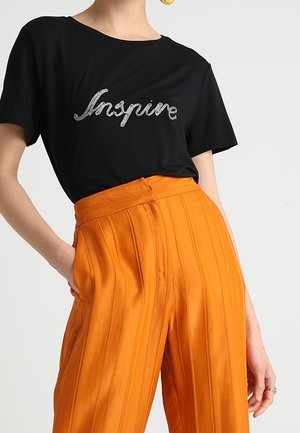 Czarna koszulka z krótkimi rękawami i srebrnym napisem "Inspire", zestawiona z wysokimi, plisowanymi pomarańczowymi spodniami wykonanymi z błyszczącego materiału.