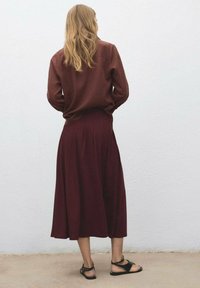 Burgundská sukně s záhyby, spárována s hnědým tričkem s dlouhým rukávem. Černé sandály na pásky celý outfit doplňují. Pozadí je bílé.